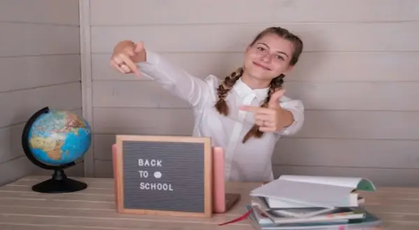 Uśmiechnięta dziewczyna z warkoczami wskazuje palcami na tabliczkę "Back to School" obok globusa i stosu książek.