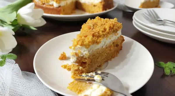 Pyszne ciasto marchewkowe z mascarpone, posypane kruszonką. Na talerzu kawałek z widelcem, obok cały tort i białe tulipany.
