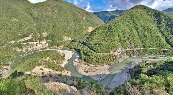 Un fiume serpeggia tra montagne verdi e scoscese, con una strada che costeggia il corso d'acqua. Il borgo di Brugnello si intravede tra la vegetazione lussureggiante.