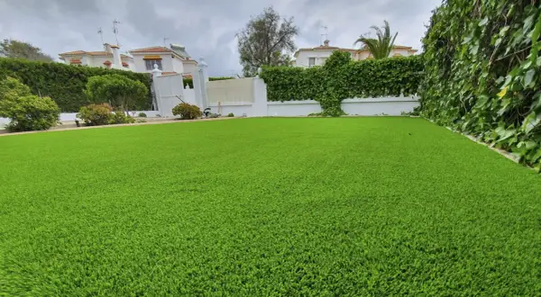 Un jardín con césped artificial impecable, delimitado por vegetación exuberante y casas blancas al fondo.
