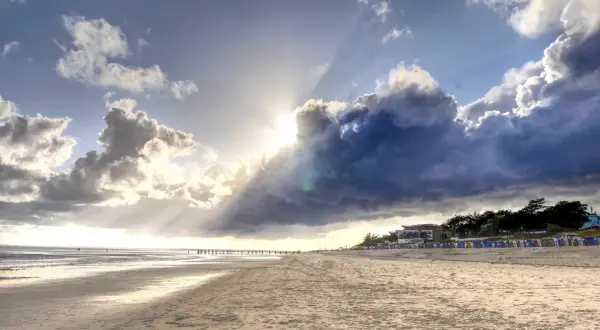 Plaża na wyspie w Niemczech. Słońce przebija się przez burzowe chmury, oświetlając piaszczysty brzeg i rzędy kolorowych budek plażowych.