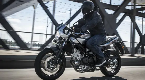 Motocyklista w kasku jedzie srebrnym cafe racerem Suzuki SV650X po moście.