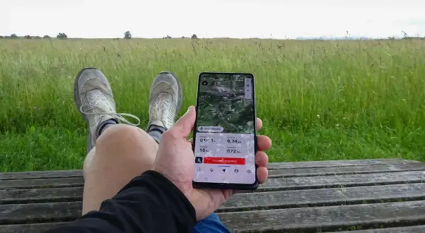 Person sitzt auf einer Holzbank, hält ein Smartphone mit einer guten Wander-App, die eine 6,74 km lange Tour mit 16 hm Anstieg und 672 hm Seehöhe anzeigt.