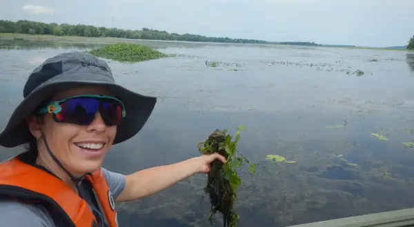 Une femme en gilet de sauvetage tient une plante aquatique, peut-être une châtaigne d'eau, dans un lac.