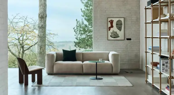 Gemütliches Wohnzimmer mit einem Sofa in millennial grey, einem Holzsessel und einem Bücherregal vor einem Fenster mit Blick ins Grüne.