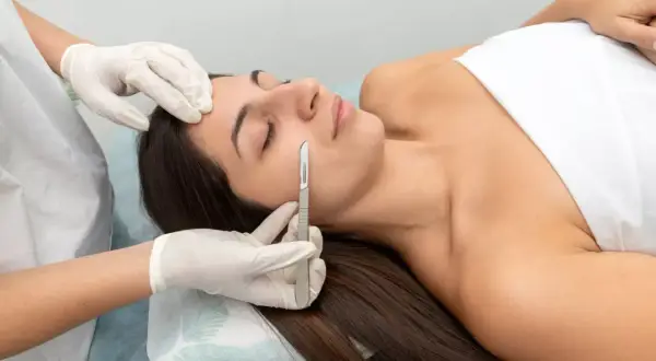 Profesional realiza dermaplaning facial a una mujer acostada, usando un bisturí para exfoliar suavemente su piel.