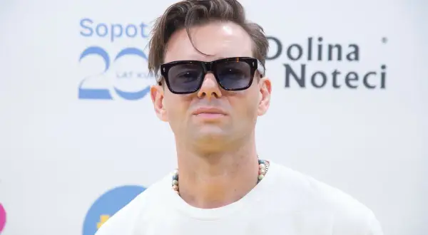 Rubens piosenkarz w okularach przeciwsłonecznych i naszyjniku z koralików, pozuje na tle napisu "Sopot 20 Lat" i "Dolina Noteci".