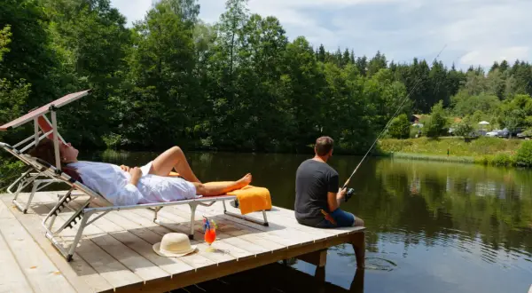 Ein Mann angelt, während eine Frau auf einer Liege entspannt. Perfekter Urlaub in Deutschland am See mit Blick auf den Wald.