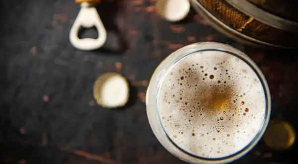 Un bicchiere di birra con una schiuma dorata e bollicine, accanto a un apribottiglie e tappi su uno sfondo scuro. La birra senza schiuma è un piacere per gli occhi.