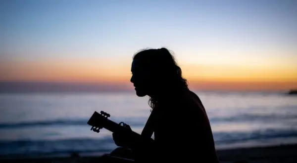 Sylwetka grającej na ukulele osoby na tle zachodzącego słońca nad morzem. Gdzie ta keja chwyty?
