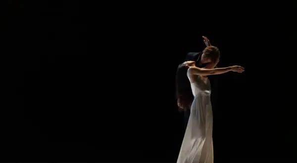 Zwei Tänzer des Tanztheater Wuppertal in inniger Umarmung auf dunkler Bühne. Die Frau im weißen Kleid neigt den Kopf zurück, der Mann beugt sich über sie.