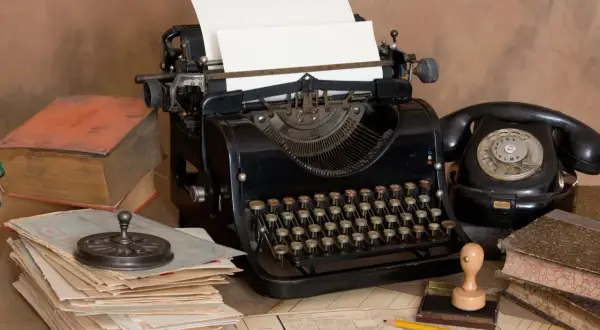 Escritorio antiguo con máquina de escribir, teléfono negro, libros y papeles. Un lápiz amarillo descansa sobre documentos, evocando la creación de dibujos con máquina de escribir.
