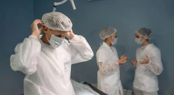 Chirurg w masce i czepku zakłada fartuch ochronny na tle sali operacyjnej.