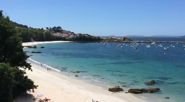 La **playa de Aguete** luce un día soleado con aguas cristalinas y barcos fondeados. Gente disfruta de la arena blanca y el mar azul.