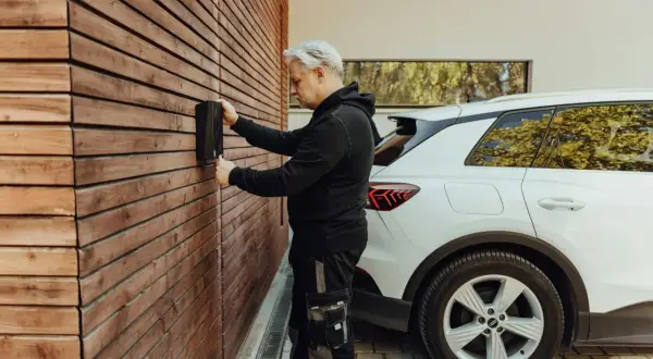 Un homme installe une borne de recharge pour voiture électrique sur un mur en bois. La question "peut on installer soi meme une borne de recharge" se pose.