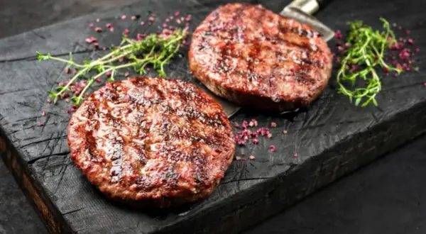 Dwa soczyste, grillowane kotlety na burgery na drewnianej desce z ziołami i różowym pieprzem.