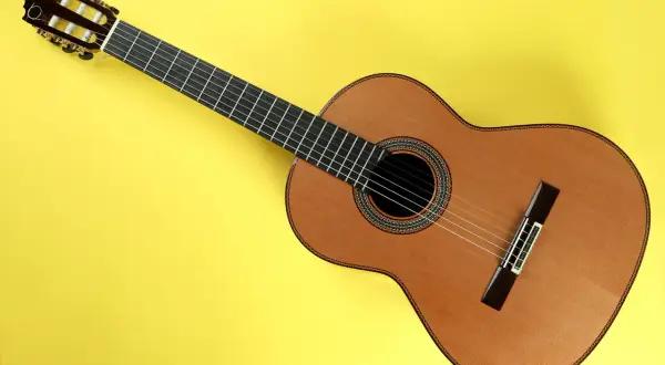 Drewniana gitara klasyczna na żółtym tle. Sześć strun, ciemna podstrunnica i ozdobne rozety wokół otworu rezonansowego. Słychać, jak brzmi gitara klasyczna.