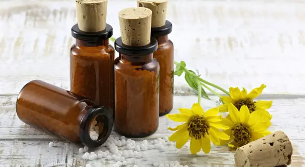 Bursztynowe buteleczki z korkami i białymi kuleczkami, symbolizujące homeopatię, obok żółtych kwiatów na drewnianym tle.