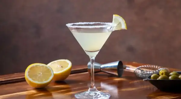 Elegancki martini bianco z solnym brzegiem i plasterkiem cytryny, obok połówki cytryny i oliwek.