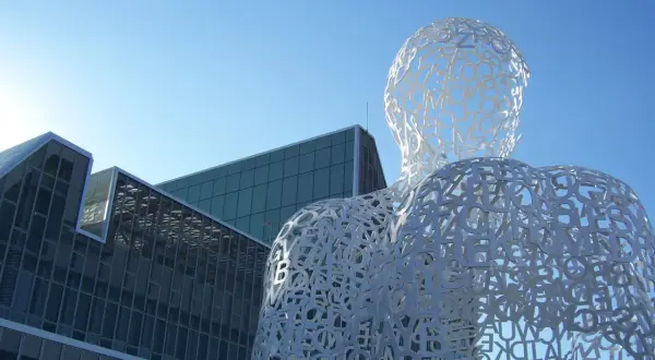 Biała, ażurowa rzeźba postaci z liter, przypominająca monumentalizm, na tle nowoczesnych budynków i błękitnego nieba.