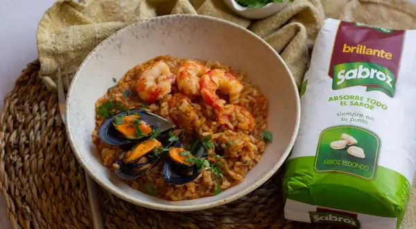 Un plato de arroz con gambas congeladas y mejillones, decorado con perejil fresco. Al lado, un paquete de arroz redondo Sabroz.