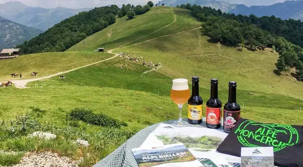 Birrificio piemontese con vista su pascoli verdi e montagne. Tre birre artigianali e un bicchiere di birra su un tavolo con materiale promozionale.