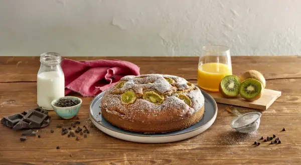 Torta kiwi e cioccolato cosparsa di zucchero a velo, con kiwi freschi, gocce di cioccolato, latte e succo d'arancia su un tavolo di legno.