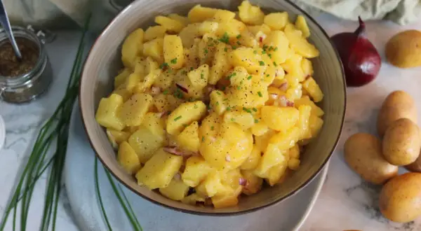 Schüssel mit leckerem Kartoffelsalat ohne Mayo, garniert mit Schnittlauch und Pfeffer. Daneben liegen frische Kartoffeln und eine rote Zwiebel.
