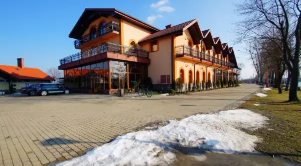Pomarańczowy budynek z balkonami i przeszklonym parterem, stojący przy drodze nad jeziorem. Roś.