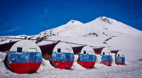 Białe, beczkowate schroniska z malowanymi flagami Rosji, zasypane śniegiem, na tle ośnieżonego Elbrusa. Wejście na szczyt.