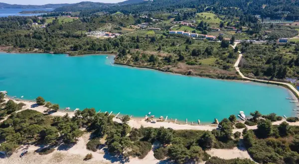 Turkusowa laguna na Kassandrze, otoczona zielonymi wzgórzami i sosnowym lasem. W oddali widać zabudowania i plażę z łodziami.
