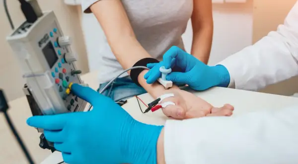 Lekarz w niebieskich rękawiczkach wykonuje próbę tężyczkową, przykładając elektrody do dłoni pacjentki i obsługując aparat.