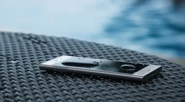 Smartfon z kroplami wody na ekranie, leżący na plecionej powierzchni. IP68 co oznacza? To wodoodporność, idealny na wakacje przy basenie.
