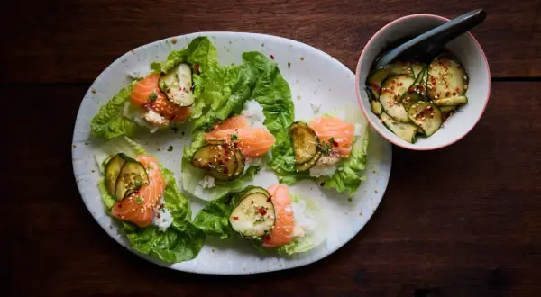 Läckra asiatiska tapas med lax, ris och gurka i salladsblad. En skål med marinerad gurka står bredvid.