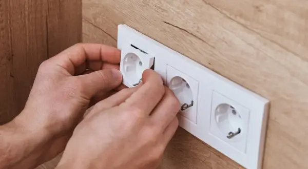 Dłonie montują białą ramkę z gniazdkami elektrycznymi na drewnianej ścianie.