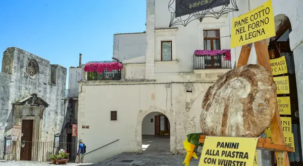 Un vicolo pittoresco a San Michele sul Gargano, con un grande pane rustico in primo piano e cartelli che pubblicizzano "Pane cotto nel forno a legna" e "Panini alla piastra".