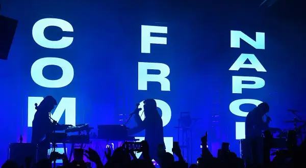Sul palco, silhouette di musicisti con tastiere e microfoni, mentre sullo sfondo campeggia la scritta "COM FROM NAPOLI" in lettere luminose.