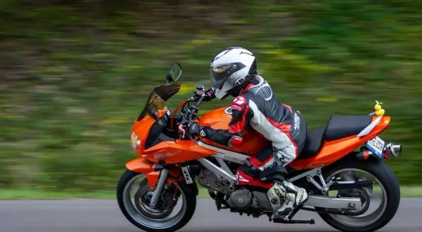 Pomarańczowy motocykl z motocyklistą w kombinezonie i kasku jedzie po drodze wśród zieleni.