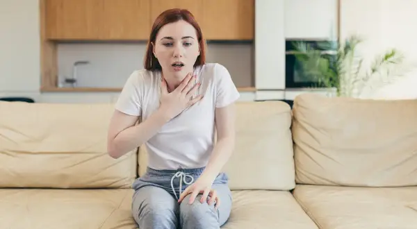 Une jeune femme assise sur un canapé, la main sur la poitrine, le souffle court. Elle semble avoir du mal à parler.