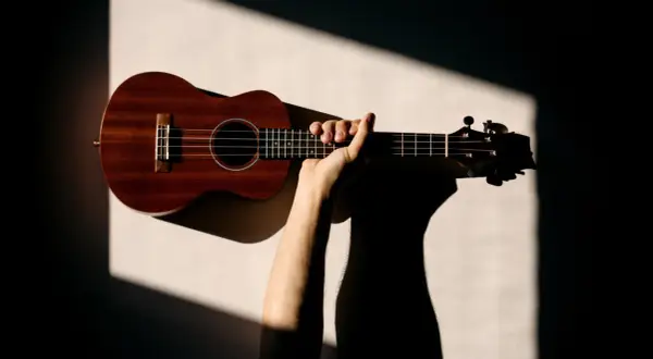 Dłoń trzymająca drewniane ukulele na jasnym tle z geometrycznymi cieniami.