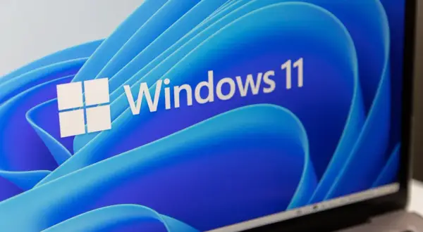 Ekran laptopa z logo Windows 11 i niebieskim, falistym tłem. System wymaga modułu TPM.