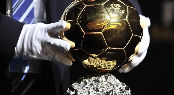 Złota Piłka FIFA Ballon d'Or na błyszczącym cokole, trzymana w białych rękawiczkach.