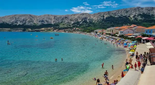 Słoneczny dzień na plaży w Baška, Krk, Chorwacja. Turkusowa woda, tłumy ludzi, góry w tle.