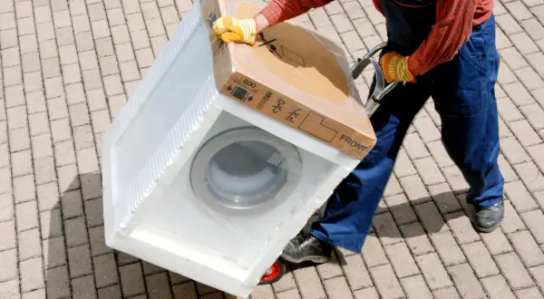 Pracownik w kombinezonie i rękawicach transportuje zapakowaną pralkę na wózku. Na kartonie widnieje napis "MAX 600 kg", co sugeruje, ile waży pralka.
