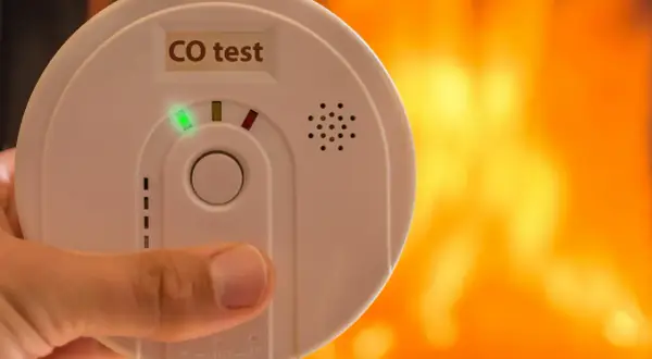Détecteur de monoxyde de carbone tenu à la main, avec un feu de cheminée en arrière-plan. Où placer détecteur monoxyde de carbone ?