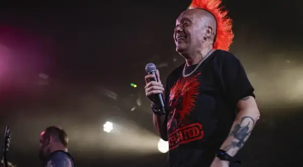 Wokalista punkowy z pomarańczowym irokezem śmieje się na scenie, trzymając mikrofon.