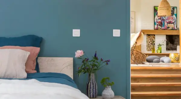 Un lit douillet avec des coussins roses et blancs sur un mur couleur bleu canard. À côté, un escalier en bois mène à un salon cosy.