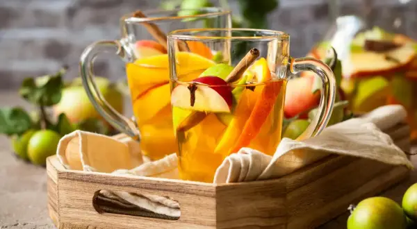 Zwei Gläser mit warmem Apfel-Zimt-Wasser, garniert mit Apfelstücken und Zimtstangen, stehen auf einem Holzteller.