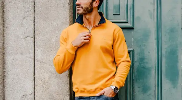 Un homme ethnic blue porte un pull jaune vif avec un col bleu marine, ajustant la fermeture éclair. Il est appuyé contre un mur en pierre.