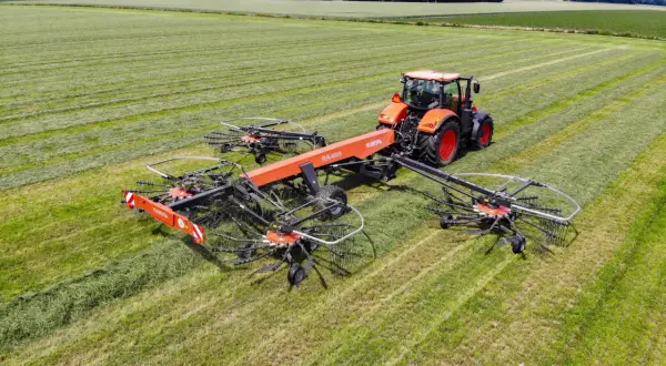 Pomarańczowy traktor zgrabnie manewruje po polu, ciągnąc szeroką zgrabiarkę do traktorka, która sprawnie formuje pokosy zielonki.